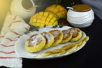 Table setting for breakfast. Curd fritters, syrniki. On a plate syrniki with bananas. Next mango and a bowl of sugar. 