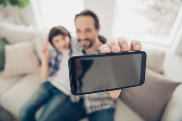 Close up photo portrait of positive relaxed nice glad daddy making selfie with his younger son on telephone sitting on comfy couch having rest free time on weekend in living room house
