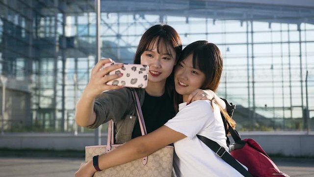 Smiling elated young asian women in modern clothes hugging and making selfie near the airport