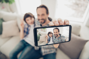 Phto portrait of handsome positive cheerful dad hugging embracing his son on couch taking selfie on telephone showing v-sign having best weekend sitting in living room indoors
