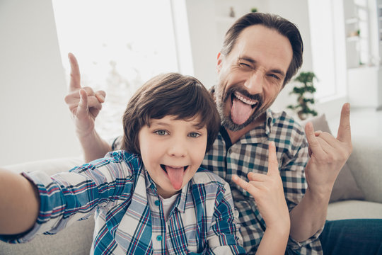 Hey there followers. Close up photo of positive cheerful handsome guy dad making hard punk symbol and his little son taking selfie on smartphone showing tongue in room indoors