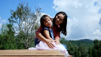 Happy mother sitting with her daughter on wooden ground over trees  with mountains background under blue sky in summer day.
