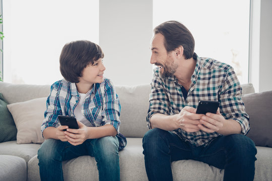 Weekend Activities Concept. Photo Of Cheerful Bearded Dad Watching Comic Video On Telephone Telling His Excited Offspring Jokes And Laughing Stories Using Telephones Sitting On Divan In Living Room