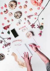 Woman hands writes in notebook on white desktop with empty screen smartphone, cup of coffee, make up cosmetics, pumpkins, fall flowers and autumn leaves. Flat lay. Top view