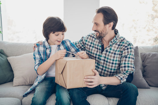 Portrait of two nice attractive lovely cheerful cheery guys sitting on sofa pre-teen son opening carton box hugging dad day birthday occasion in light white modern interior house living-room