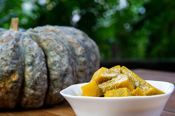 Yummy pumpkin and sweet coconut milk in white bowl, popular Thai dessert and Ripe fresh Thai pumpkin on wooden table with green nature blurred background.