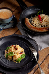Delicious lunch - spaghetti, meat sausage, chili pepper in frying pan, black round plate with Italian pasta with vegetables, basil leaves, vintage cutlery, fork, blue cup americano coffee
