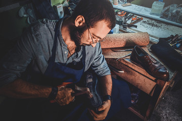 Diligent cobbler is working on shoe sole using hammer at his dark workplace.