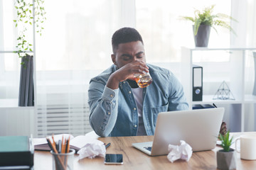 Tired african american employee drinking alcohol at workplace