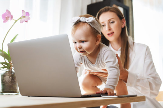 Young mother with baby girl sitting at home in front of computer and watching funny videos for children. Mom showing her toddler daughter modern digital gadgets using for work and education online.