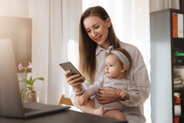Little girl sitting on mother's knees and looking at screen of mom's smartphone. Young woman and...