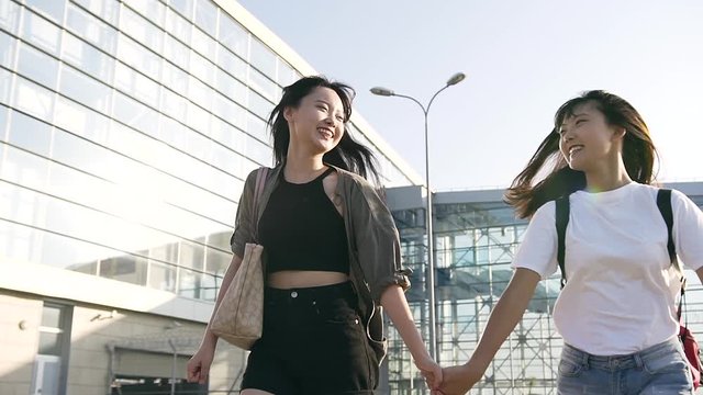 Front view of jolly good-looking young asian girls in trendy clothes which holding by hands and running near the modern airport