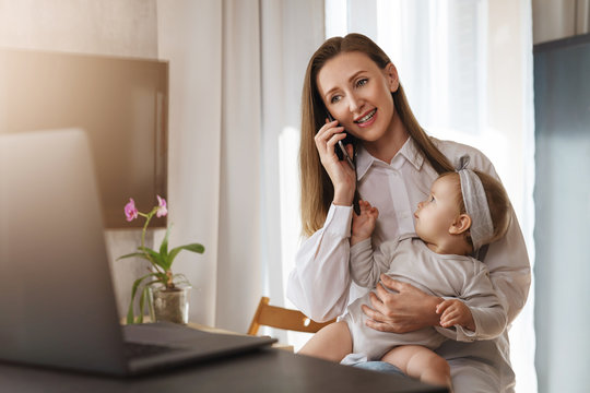 Young woman making phone call on her mobile, baby daughter sitting on knees and watching at mom. Mother working on laptop remotely while taking care of child at home. Freelance career for young moms
