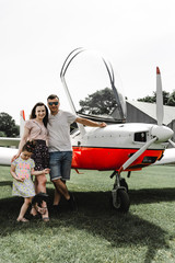 Happy family having fun on the green grass near airplane. Beautiful couple with little daughter posing on the aircraft. Walk at airport