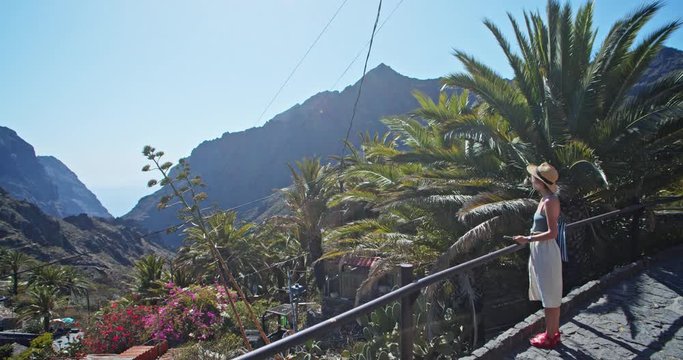 Inspired young beautiful woman observing Masca gorge valley panorama on viewpoint. Zoom drone shot of spectacular village nature scenery and girl tourist. Santa Cruz de Tenerife, Spain.