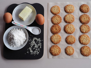 Recipe delicious sesame cookies in detail. Flour, eggs, sesame seeds, butter in plates, a spoonful of sugar on a dark stand and sesame cookies on parchment.