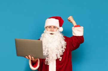 Fototapeta na wymiar Joyful Santa Claus winner with laptop in hand isolated on blue background, looks into camera and rejoices. Happy guy in santa suit rejoices in victory, holds laptop. New Year concept.