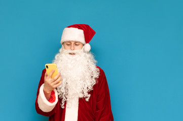 Fototapeta na wymiar Smiling young man in santa suit standing on blue background and using smartphone, isolated. Santa teenager with smartphone isolated on blue background. Christmas concept.