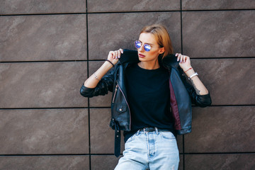 Girl wearing t-shirt, glasses and leather jacket posing against street , urban clothing style....