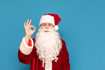 Fototapeta na wymiar Happy young Santa Claus with a beard and in a red hat stands on a blue background, shows his finger gesture Ok and looks into the camera. Guy in the santa suit shows the sign OK and smiles.