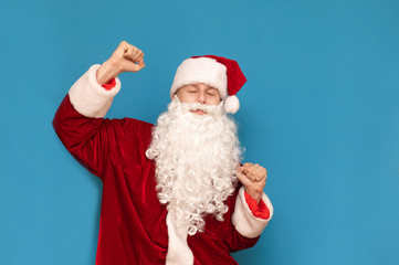 Fototapeta na wymiar Cheerful young man in costume of santa claus dancing on blue background with closed eyes. Happy guy in santa costume having fun, dancing, isolated. Christmas party.