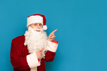 Fototapeta na wymiar Funny guy in santa suit isolated on blue background, pointing to the empty space, looking into the camera and smiling. Positive Santa for Christmas and copyspace