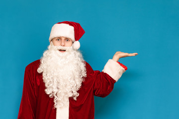 Fototapeta na wymiar Funny young Santa Claus in hat and white beard, looks at camera in amazement and shows his hand on copyspace. Isolated on blue background. Santa is pointing to an empty space. Background