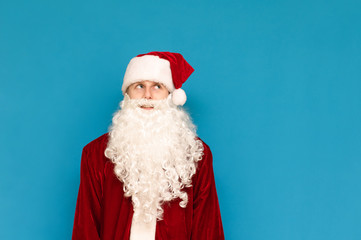 Funny guy in santa costume isolated on blue background, stands on looks aside with a smile on his face. Cheerful teenager in the image of Santa at a Christmas party. Studio portrait.