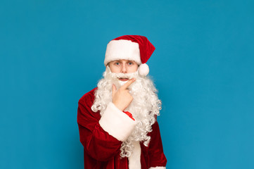 Portrait of a funny pensive teenager in Santa Claus costume, looking intently into the camera, isolated on blue background. Young Santa in Costume looks in camera and thinks.Christmas and New Year