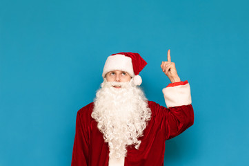 Fototapeta na wymiar Cute young Santa Claus isolated on a blue background, looking and pointing his finger up at a blank space. Santa's cheerful guy points his finger at Copyspace. Christmas party concept.