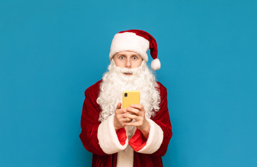 Shocked young man in santa claus christmas costume, standing with smartphone in hands and with surprised face looks in ccamera on blue background.Surprised Santa with smartphone in hand,isolated