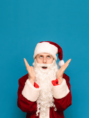 Portrait of a frightened guy in a suit of Santa Claus on a blue background, shocked with raised hands to face looking up at a blank spot. Shocked young Santa looking at copy space. Vertical