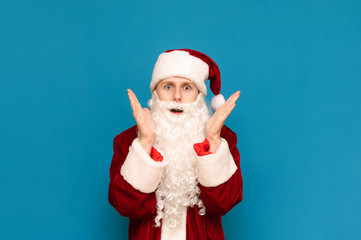 Portrait of a frightened young Santa Claus on a blue background, looks shocked face to camera and raised his hands to face in fear. Isolated. Copy space