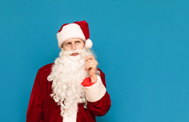 Fototapeta na wymiar Closeup portrait of a pensive santa on a blue background, looking away at an empty space and thinking. Young guy in santa suit isolated on blue background. christmas concept.