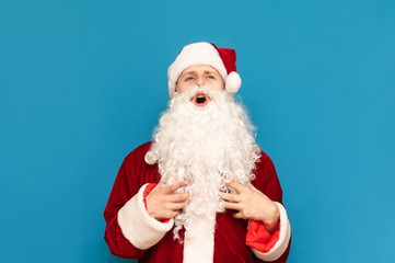 Funny young santa claus with beard and hat laughs against blue background and looks up. Photo of funny guy in santa costume smiling. Isolated.