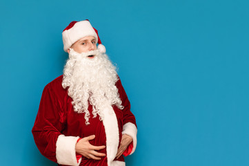 Funny young man in Santa Claus costume and big belly posing on camera against blue background. Funny young santa holds her belly and looks into the camera.