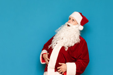 Funny young Santa with beard, hat and big belly isolated on blue background, looking away. A teenager dressed himself in a Santa Claus costume and poses for the camera. Christmas concept.
