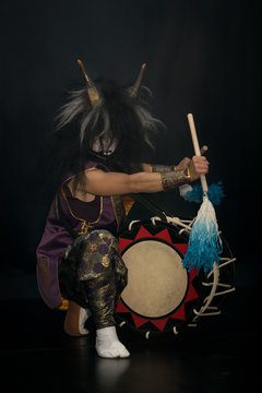 Taiko drummer in a wig and a demon mask on stage squarts with drumstick and drum on a black background. Demon from Japanese mythology.