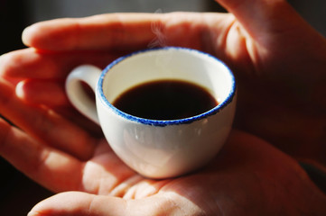 Close-up of Woman's hands holding a tiny white ceramic cup of hot black coffee. Selective focus