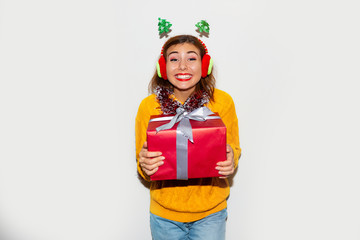  Portrait of a smiling young girl in yellow knitted sweater showing gift box and looking at camera isolated over gray background. Holding Christmas gift in red box with ribbon. 