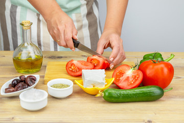 Woman cuts tomatoes for Greek salad. Ingredients for Greek Salad on wooden background. Healthy food, Mediterranean diet. Girl cooking Greek Salad with fresh vegetables.