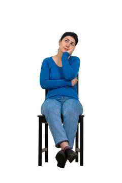 Full Length Portrait Of Casual Young Woman Seated On A Chair Keeps Hand Under Chin Looking Up Thoughtful Isolated Over White Background With Copy Space.