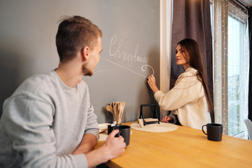 Young married couple in casual clothes, man and woman, sitting at home in the kitchen at the table. Draw Merry Christmas with chalk on the wall.