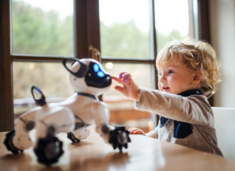 A cute toddler boy standing indoors at home, playing with robotic dog.