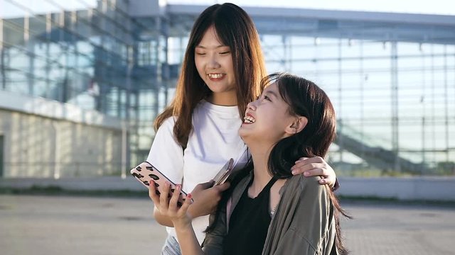 Two lovely exuberant young asian girls smiling from revision of funny photos on phone near modern building