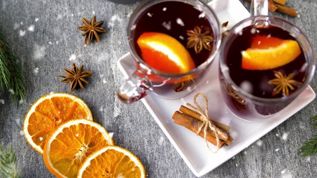 christmas and seasonal drinks concept - snowing over glasses of hot mulled wine with orange slices, aromatic spices and fir branch on grey background