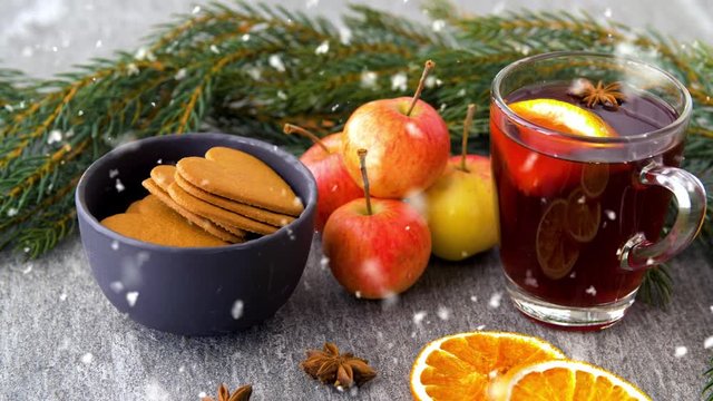 christmas and seasonal treats concept - snowing over glass of hot mulled wine with orange slice, gingerbread cookies, apples and fir branch on grey background
