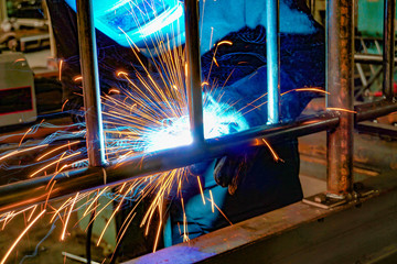 A welder at work in a workshop produces metal structures. Sparks from welding fly around.