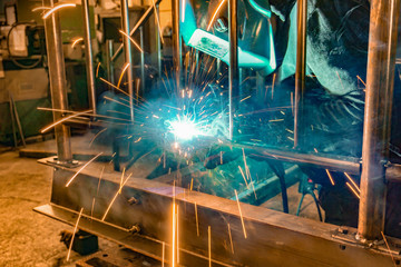 A welder at work in a workshop produces metal structures. Sparks from welding fly around.