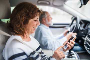 Happy senior couple with smartphone sitting in car, going on trip.